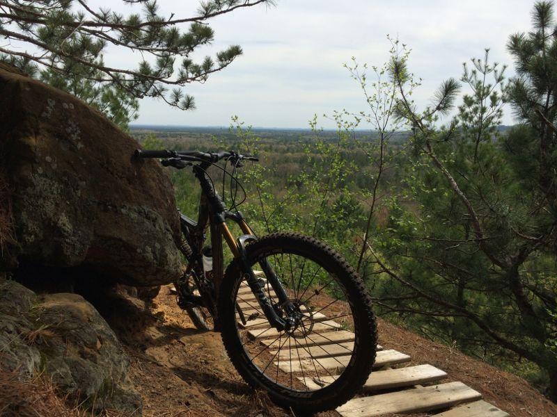 A mountain bike rests beside a large rock on a wooded trail, overlooking a vast landscape of trees and hills in the background under a cloudy sky. Levis Mounds mountain bike trail.