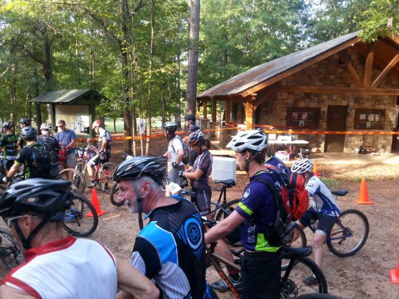 A group of mountain bikers gathered near a wooden shelter in a forested area, preparing for a biking event. Participants are wearing helmets and cycling jerseys, with some bikes parked nearby. The scene includes orange cones and a backdrop of trees, creating an outdoor atmosphere for the activity. Camp Thunder mountain bike trail.