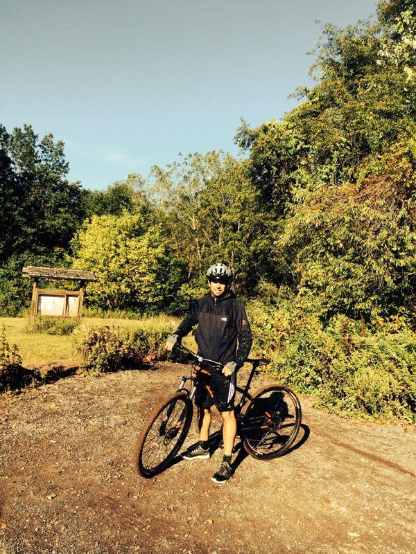 A person wearing a helmet and athletic clothing stands beside a mountain bike on a gravel path, surrounded by greenery. In the background, there is a wooden signpost near a grassy area. The sky is clear and blue, indicating a bright day. Six Mile Run mountain bike trail.