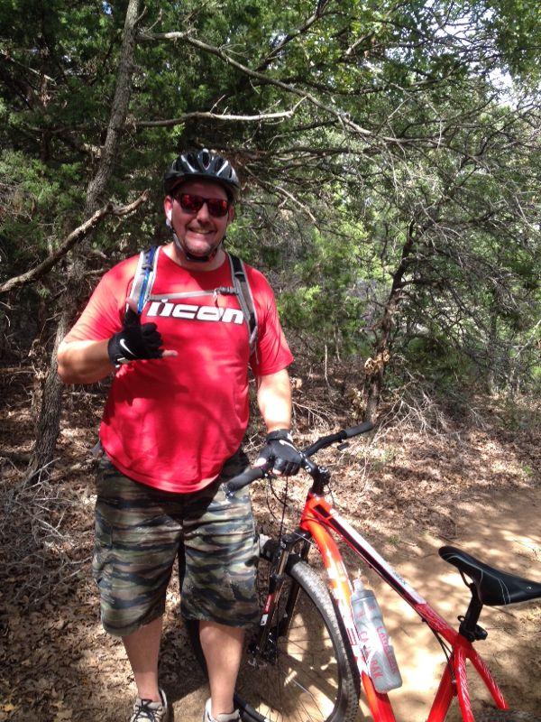 A man wearing a red t-shirt and camo shorts stands next to a red mountain bike on a wooded trail, giving a thumbs up. He is wearing a bike helmet and sunglasses, and he has a backpack on his back. The surrounding area consists of green trees and underbrush. McMurtry Trail mountain bike trail.
