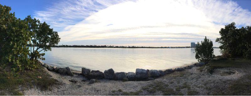A panoramic view of a serene waterfront scene featuring calm waters reflecting the sky. Lush green foliage lines the shore, with scattered rocks along the water's edge. The sky above is partly cloudy, showcasing a mix of blue and soft white clouds. In the distance, a hint of urban structures is visible along the horizon. Oleta River State Park mountain bike trail.