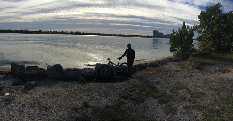 A person stands next to a bike on a rocky shore by a calm body of water, with trees and buildings in the background. The sky is filled with clouds and has a soft, warm light reflecting on the water's surface. Oleta River State Park mountain bike trail.