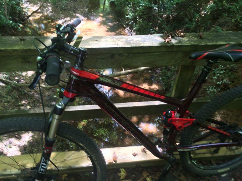 A mountain bike resting on a wooden bridge above a small stream, surrounded by lush greenery. The bike features a reddish frame with black and red accents, showcasing its various components like the handlebars and tires. In the background, the creek flows gently through a tranquil wooded area. Dauset Trails Nature Center mountain bike trail.