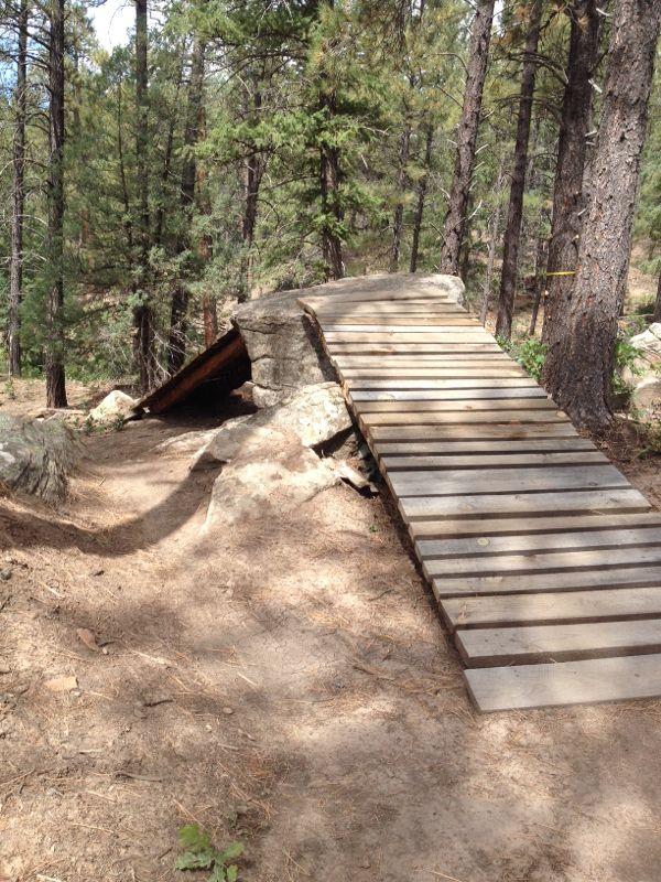 A wooden ramp built over rocky terrain, leading to a platform made of stone, surrounded by dense trees in a forested area. The ground is covered with pine needles and dirt, creating a natural hiking or biking path. Twin Buttes mountain bike trail.