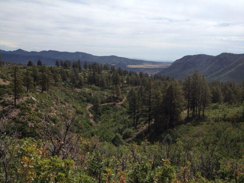 A scenic view of a mountainous landscape featuring rolling hills covered with evergreen trees and patches of greenery. The sky is partly cloudy, and distant mountains are visible on the horizon, creating a peaceful natural setting. Twin Buttes mountain bike trail.