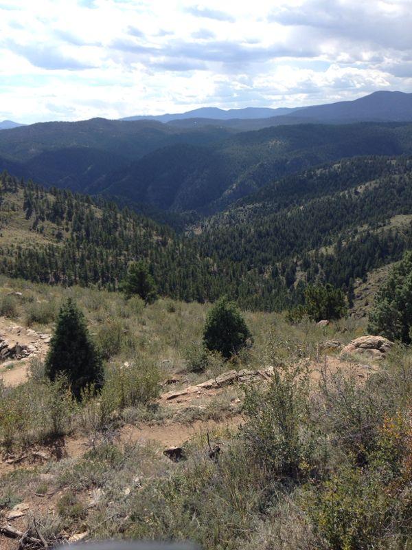 A panoramic view of a mountainous landscape featuring rolling hills covered in green vegetation, sparse trees, and rocky terrain under a partly cloudy sky. The scene showcases a deep valley nestled between the mountains, creating a picturesque natural setting. Centennial Cone Park mountain bike trail.