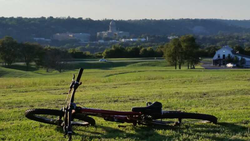 A red mountain bike lying on a grassy field with a view of a city skyline in the background, featuring a prominent dome-shaped building and rolling hills under a clear sky. Capital View mountain bike trail.