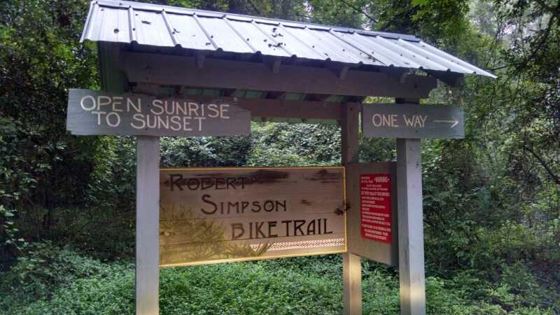 Signpost marking the entrance to the Robert Simpson Bike Trail, surrounded by lush greenery. The sign indicates the trail is open from sunrise to sunset and features a one-way arrow. Robert Simpson bike trail mountain bike trail.