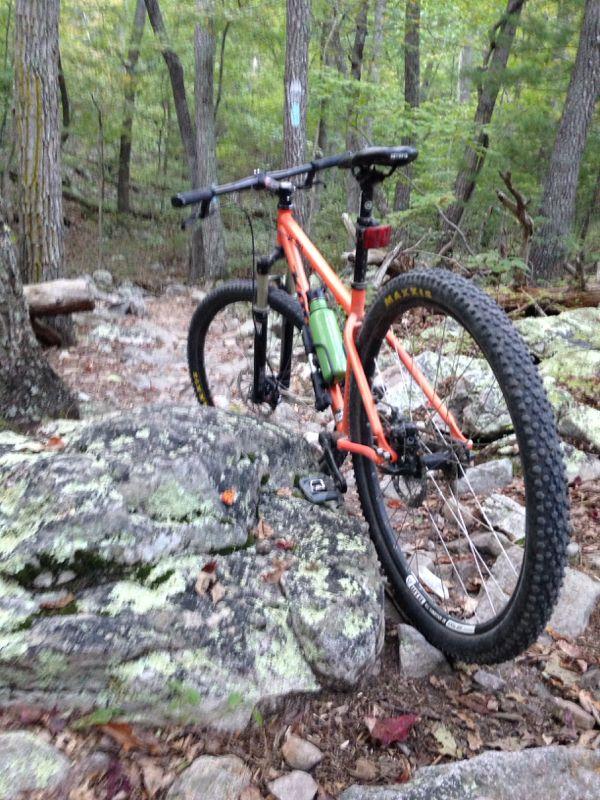 An orange mountain bike parked on rocky terrain in a forested area, surrounded by trees and foliage. The bike features thick tires suitable for off-road biking and has a water bottle mounted on the frame. Elizabeth Furnace mountain bike trail.