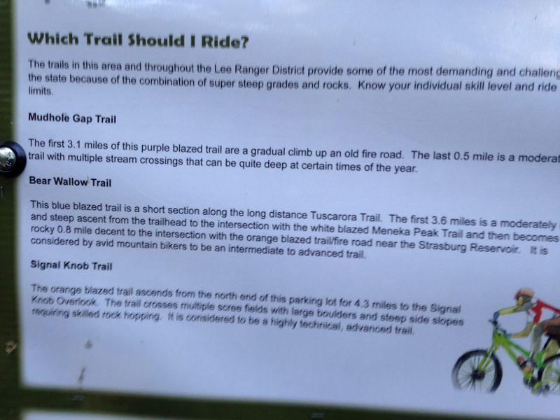 Image of a trail information sign detailing three different biking trails in the Lee Ranger District. The sign describes Mudhole Gap Trail, Bear Wallow Trail, and Signal Knob Trail, highlighting their distances, difficulty levels, and specific features like stream crossings and rocky terrain. An illustration of a cyclist is included on the side. Elizabeth Furnace mountain bike trail.