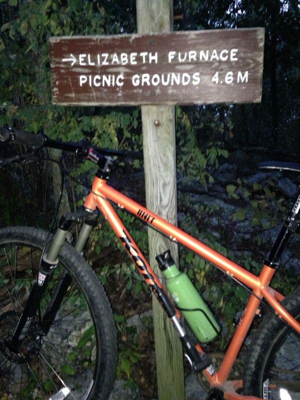 A mountain bike is leaning against a wooden signpost that indicates the direction to Elizabeth Furnace Picnic Grounds, located 4.6 miles away. The bike is orange with a green water bottle attached, and the surrounding area features dense foliage and rocks, suggesting a natural setting. Elizabeth Furnace mountain bike trail.