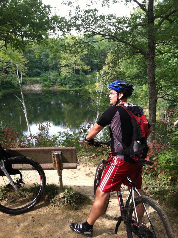 A cyclist in a black shirt and red shorts stands beside his mountain bike, looking out over a serene pond surrounded by lush greenery. A wooden bench is visible nearby, indicating a resting spot along a nature trail. Kickapoo mountain bike trail.