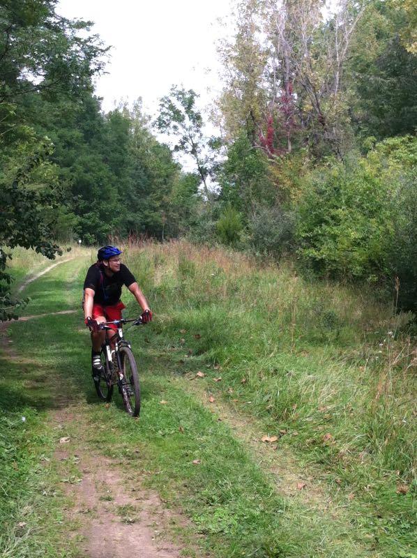 A person riding a mountain bike on a grassy trail surrounded by trees and greenery. The cyclist is wearing a helmet and athletic clothing, navigating a winding path through a natural landscape. Kickapoo mountain bike trail.