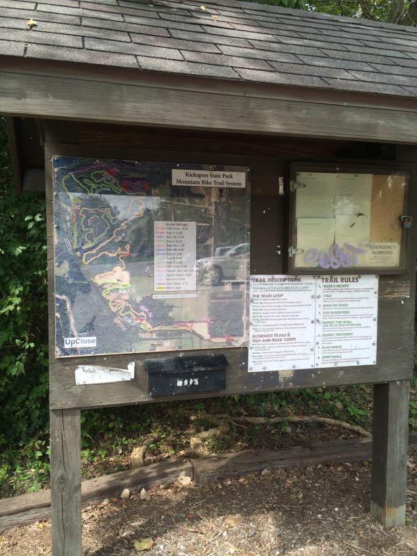 Signboard displaying a map and information for the Kickapoo State Park Mountain Bike Trail System, including trail descriptions, rules, and a map box. The board is surrounded by greenery and a gravel path is visible in the foreground. Kickapoo mountain bike trail.