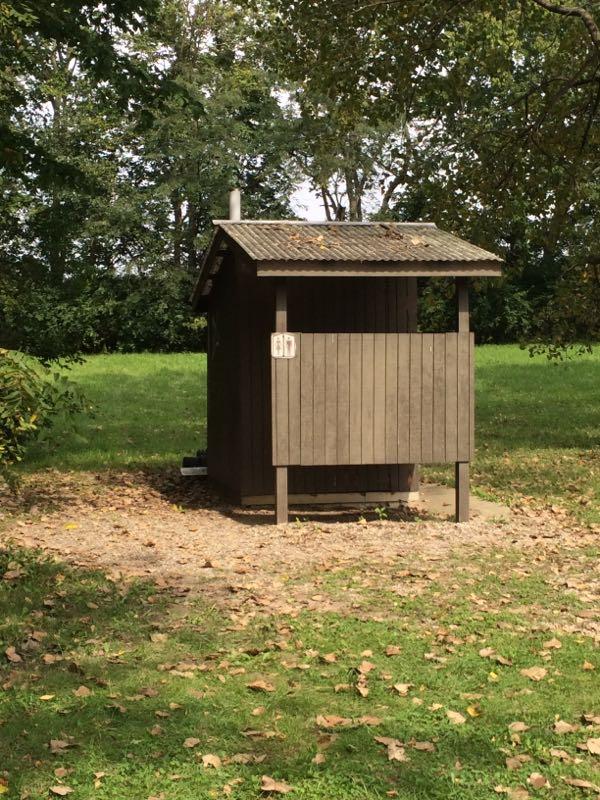 A small wooden outdoor restroom structure located in a grassy area surrounded by trees. The building has a slanted roof, a wooden door with a number plaque, and is partially shaded by nearby foliage. Leaves scattered on the ground indicate it is autumn. Kickapoo mountain bike trail.