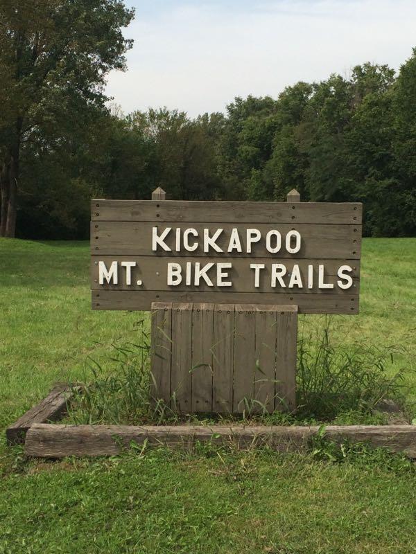A wooden sign that reads "KICKAPOO MT. BIKE TRAILS" stands in a grassy area surrounded by trees under a clear sky. Kickapoo mountain bike trail.