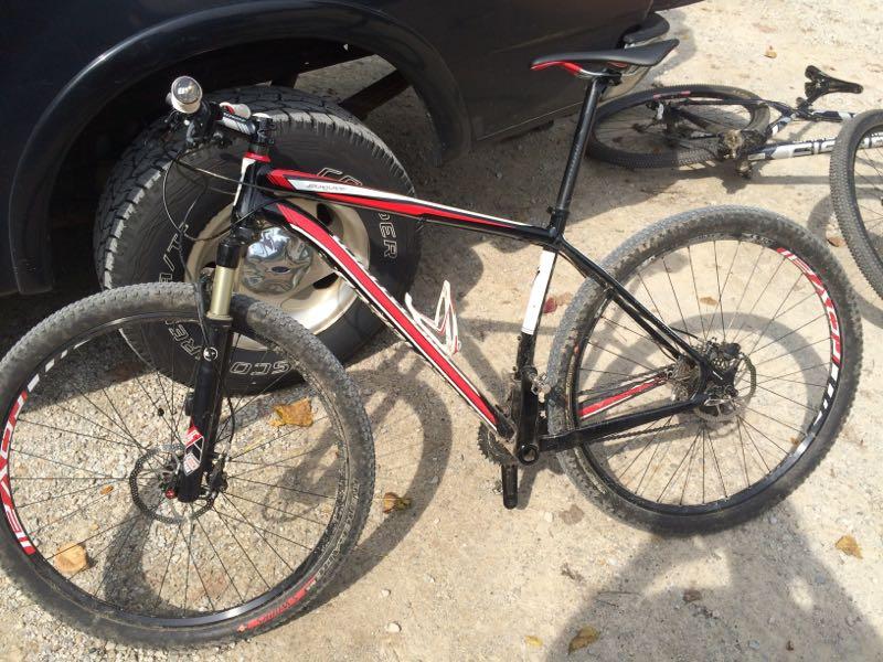A black and red mountain bike with thick tires is parked on a gravel surface. In the background, a large tire from a vehicle is visible, along with another bike partially obscured. The bike showcases a lightweight frame and features a front suspension fork, indicating it is designed for off-road cycling. Kickapoo mountain bike trail.