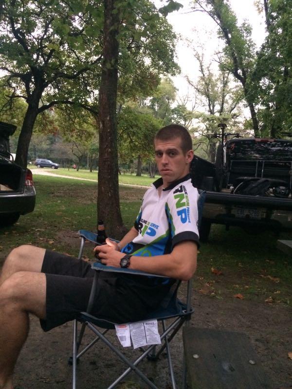 A young man sitting on a folding chair in a park, wearing a cycling jersey and shorts, with a serious expression. He is holding a drink and is surrounded by trees and parked vehicles in the background. Kickapoo mountain bike trail.
