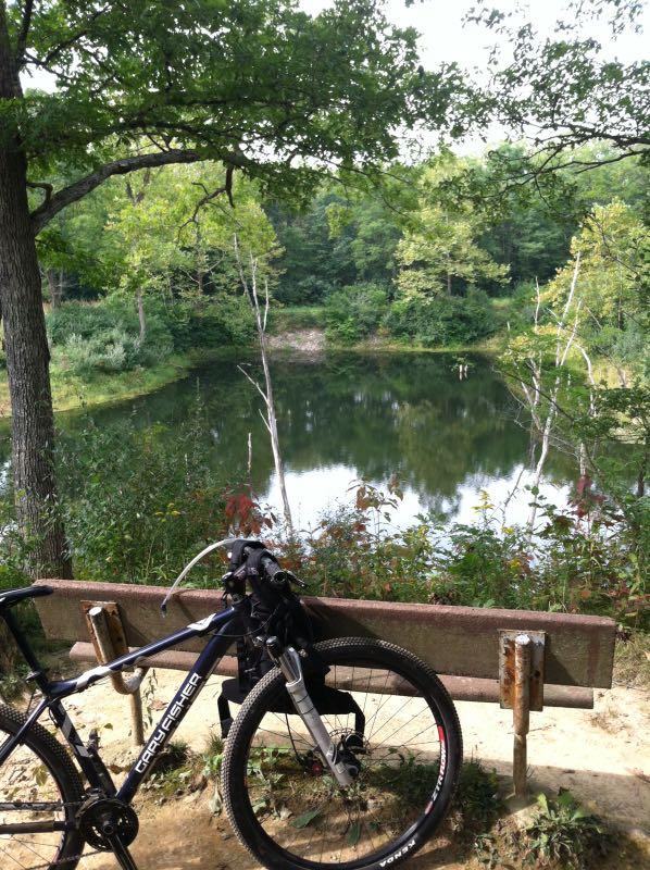 A mountain bike parked next to a wooden bench overlooking a tranquil pond surrounded by lush greenery and trees. The scene captures a peaceful natural setting, perfect for outdoor activities like biking and enjoying nature. Kickapoo mountain bike trail.