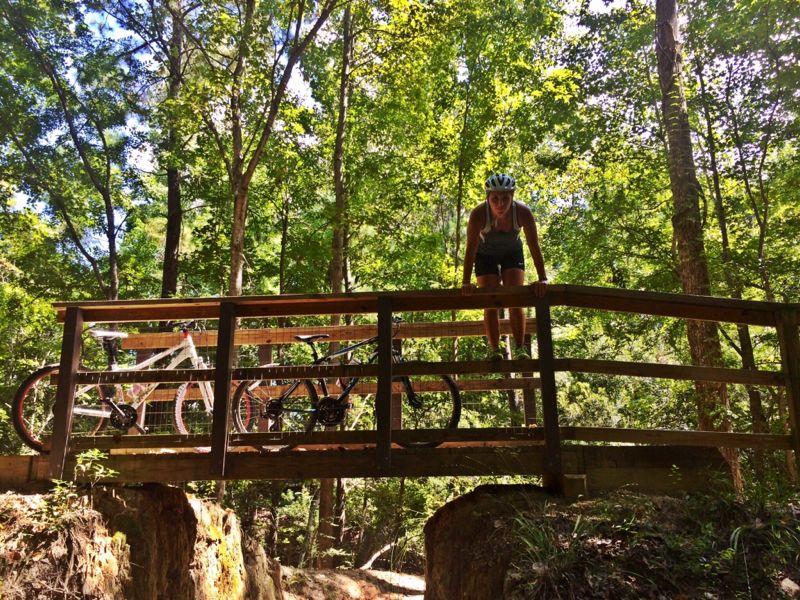 A person wearing a helmet and athletic clothing is balancing on a wooden bridge in a forested area. Two bicycles are parked on the bridge beside them, surrounded by lush green trees and dappled sunlight filtering through the foliage. Wannamaker Park mountain bike trail.