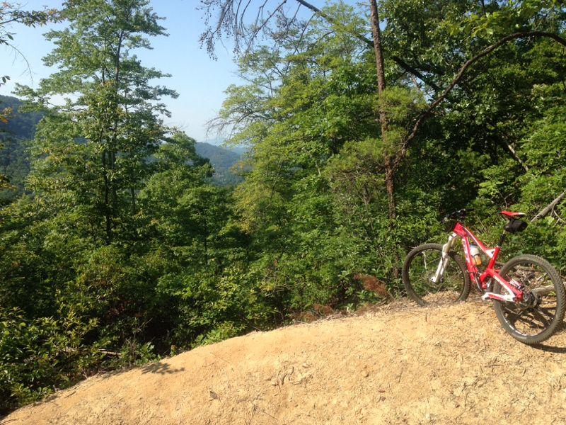 A mountain bike resting on a sandy ledge, surrounded by lush green trees and a scenic view of distant hills under a clear sky. Buffalo Creek Park: The Head, The Heart &amp; The Tail (HHT) mountain bike trail.
