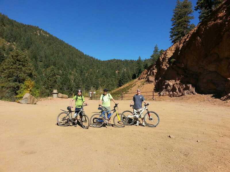 Three mountain bikers wearing helmets and casual clothing pose with their bicycles on a dirt trail surrounded by trees and hills. The sky is clear and blue, indicating a sunny day. They are situated in a natural outdoor setting with rocky terrain in the background. Captain Jack's mountain bike trail.