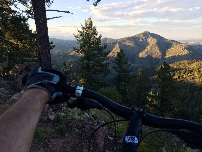 A mountain biker's hand gripping the handlebars of a mountain bike, with a scenic view of rugged mountains and trees in the background under a partly cloudy sky. Mount Falcon Park mountain bike trail.