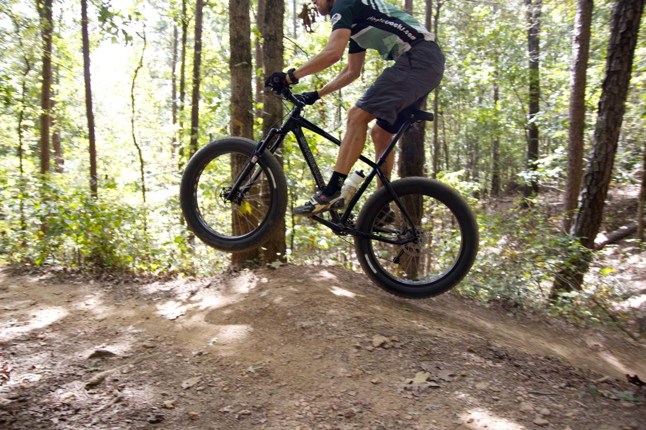Motobecane NightTrain Bullet: A mountain biker performing a jump on a dirt trail surrounded by trees. The rider is in mid-air, showcasing an action shot with focus on the bike's broad tires and the natural forest backdrop.