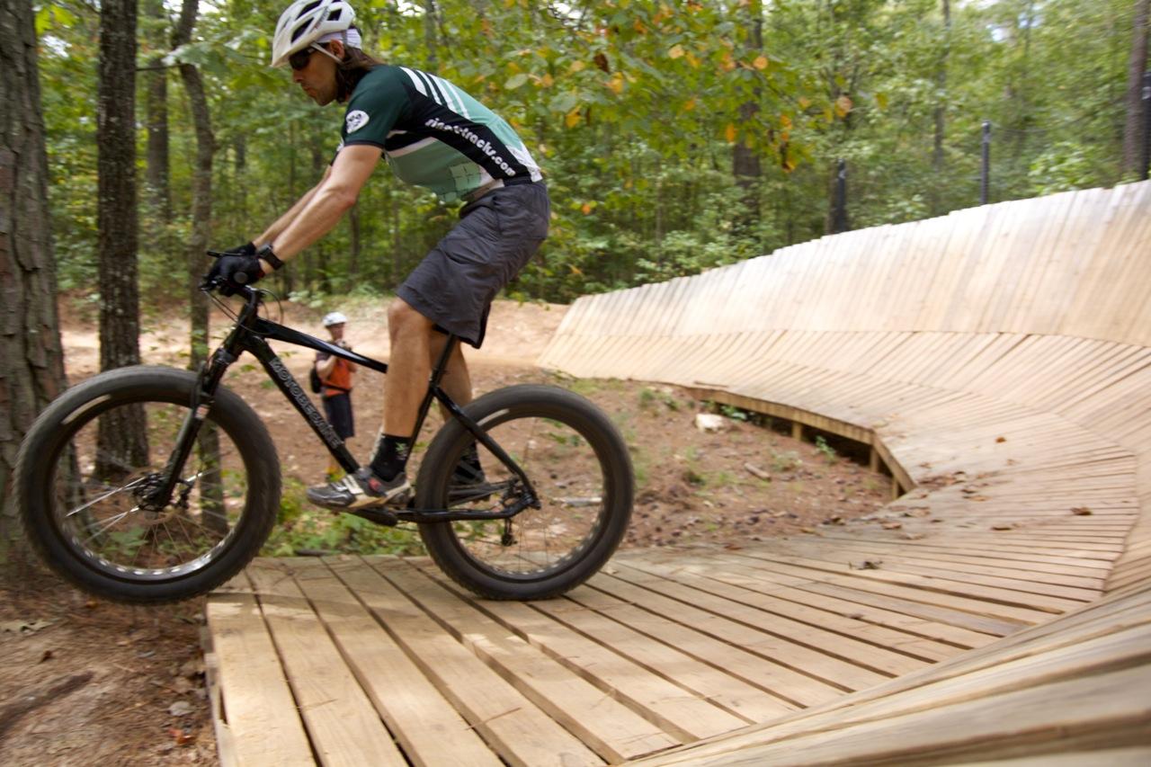 Motobecane NightTrain Bullet: A person riding a mountain bike on a wooden ramp in a forested area, with trees in the background. The cyclist is focused on navigating the curved structure, while a person in the distance watches. The ramp features a smooth, sloped design intended for biking.