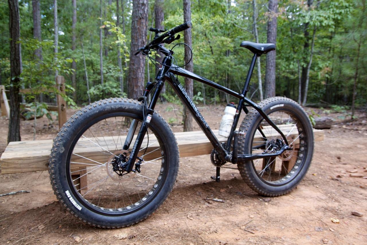 Motobecane NightTrain Bullet: A black fat tire bike is parked on a wooden bench in a wooded area. The bike features wide tires designed for off-road terrain, and a water bottle is mounted on the frame. Surrounding the bike are tall trees and natural forest ground.