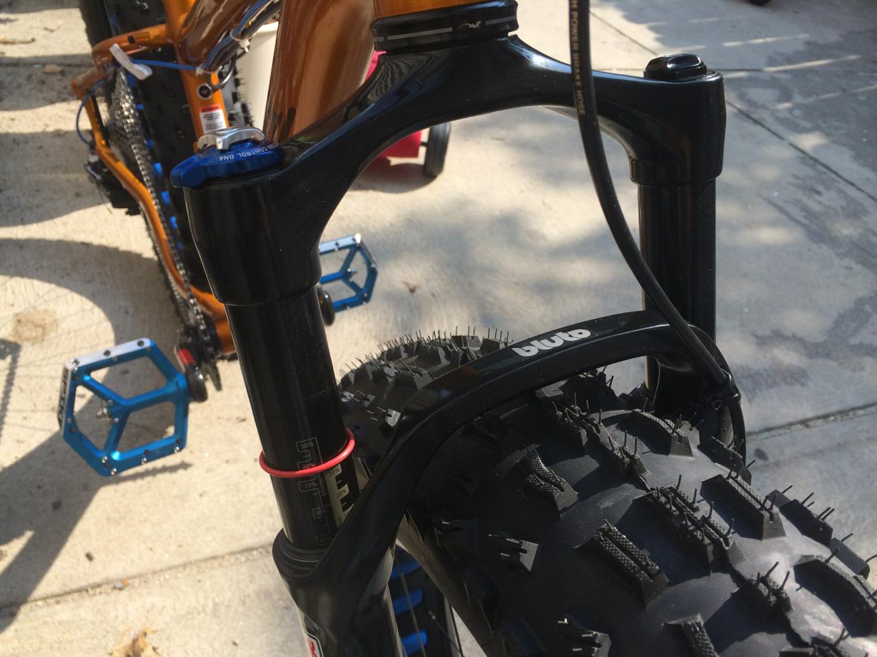 Salsa Mukluk: Close-up of a mountain bike's front fork and tire, featuring a thick, knobby tire and a black suspension fork with adjustment dials. The background shows the frame of the bike in an outdoor setting.