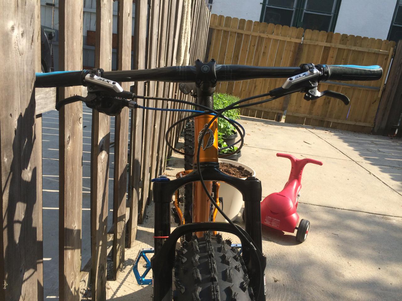Salsa Mukluk: A view from behind the handlebars of a mountain bike, showcasing the grips and brake levers. In the background, there is a wooden fence and a small red toy vehicle on a concrete surface. Pedals and a potted plant can also be seen to the side.