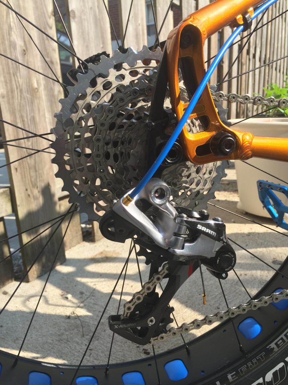 Salsa Mukluk: Close-up of a bicycle's rear derailleur and cassette, showcasing a shiny silver SRAM derailleur and a detailed view of the sprocket gear. The bike has an orange frame highlighted by blue cable housing, with a background of wooden fencing and a hint of greenery. The image captures the intricate mechanics of the bike's drivetrain components.