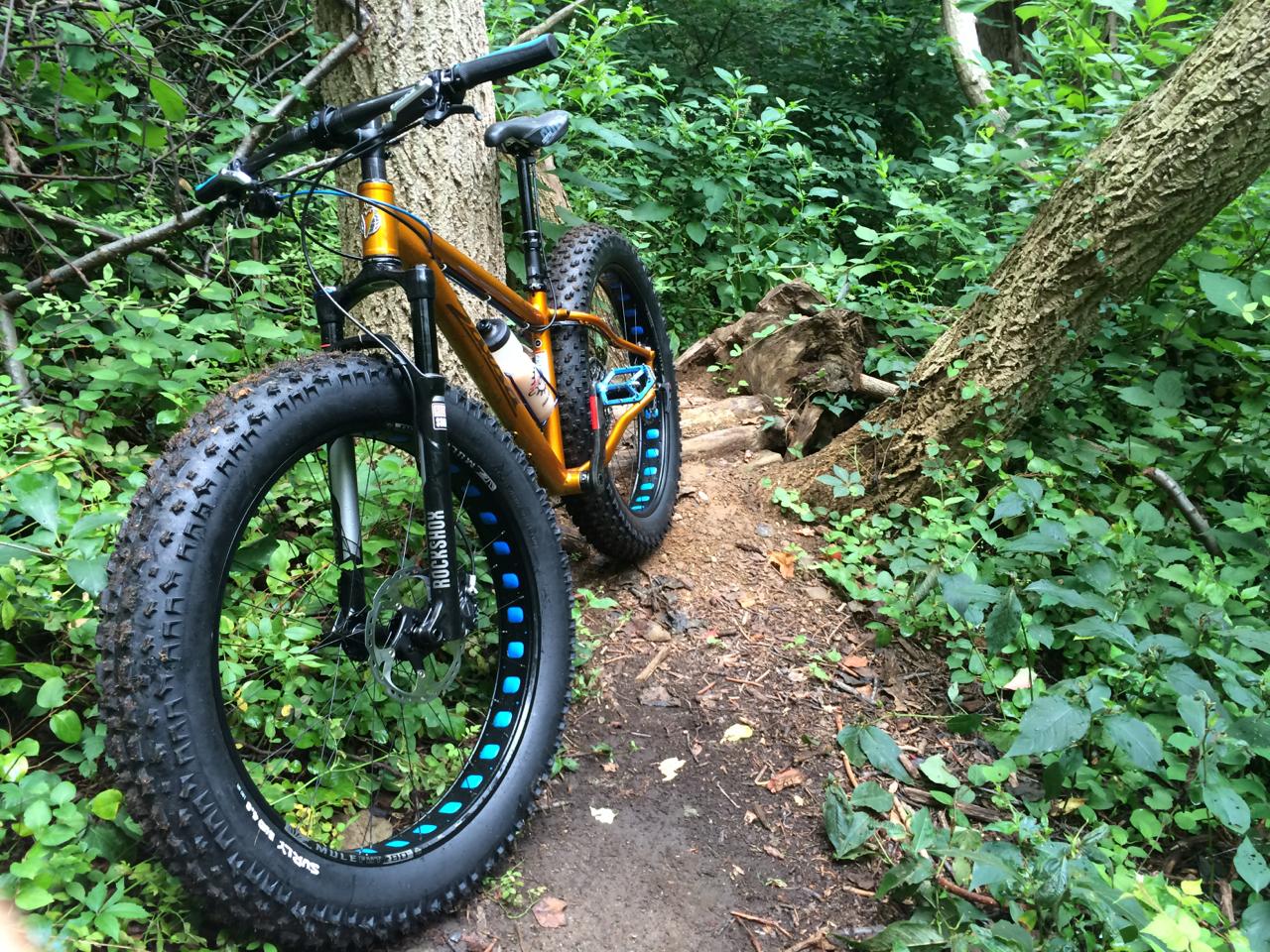 Salsa Mukluk: A mountain bike with wide tires is positioned on a narrow dirt trail surrounded by dense green foliage and trees. The bike has an orange frame with blue accents and is partially obscured by leaves and branches.