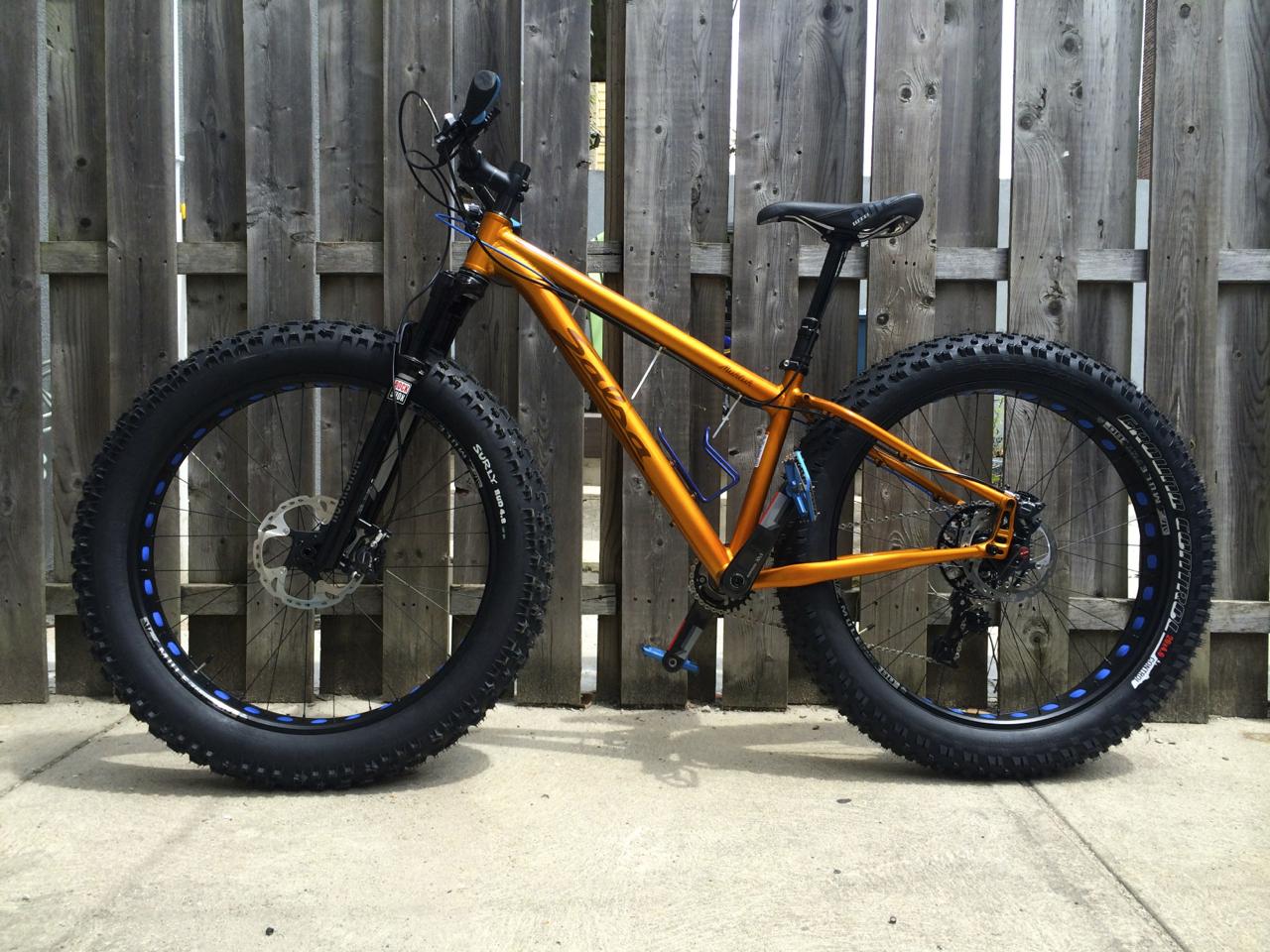 Salsa Mukluk: A bright orange fat tire bike is leaning against a wooden fence. It features thick, knobby tires and a sturdy frame, designed for handling rough terrains. The bike includes a black front fork, disc brakes, and a saddle seat, showcasing a sleek and modern design. The ground is concrete, providing a clear view of the bike's features.