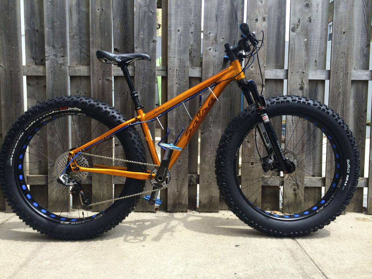 Salsa Mukluk: A bright orange fat bike with large, knobby tires stands against a wooden fence. The bike features a sleek design with a black seat, handlebars, and a gear system visible on the rear wheel. The tires are prominently marked, and the bike's frame showcases a combination of orange and blue accents. The ground surface is concrete, enhancing the bike's rugged appearance.