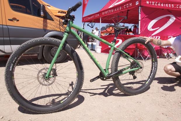 Niner ROS 9 Plus: A green mountain bike with oversized tires is displayed on a dirt surface, with a banner and van in the background from the Niner Bikes brand. A person is crouching next to the bike, showcasing its features.