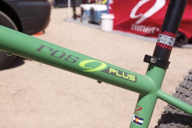 Niner ROS 9 Plus: Close-up of a green bicycle frame featuring the text "ROS 9 PLUS," with a visible Colorado flag sticker. The background shows a blurred campsite setup with a red banner and various equipment.