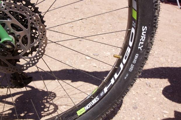 Niner ROS 9 Plus: Close-up of a bicycle wheel showing a portion of the tire and disc brake assembly. The wheel is labeled with the brand "Surly" and features a "Nobles" tire. The background is a dirt surface, suggestive of an outdoor biking environment.