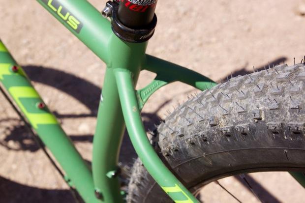 Niner ROS 9 Plus: Close-up view of a green bicycle frame and a thick, textured tire, set against a sandy background. The bicycle features a modern design with visible components like the wheel and suspension.