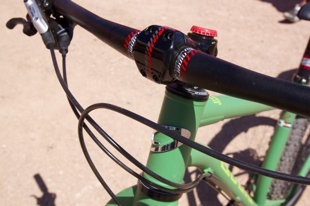 Niner ROS 9 Plus: Close-up view of a bicycle handlebar and stem, featuring a green frame, black handlebars with red accents, and visible brake and gear cables on a gravel surface.