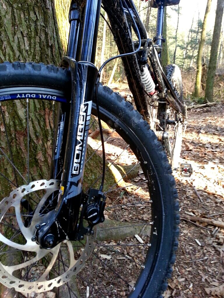 Marin 2007 Marin Quake 7.1: Close-up view of a mountain bike fork and front wheel, showcasing the disc brake system and tire tread. The bike is positioned next to a tree in a forested area, with muddy and earthy elements indicating recent outdoor use.