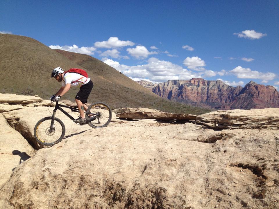 A mountain biker navigating a rocky terrain with distant mountains and a blue sky in the background, showcasing an adventurous outdoor setting. Guacamole Mesa mountain bike trail.