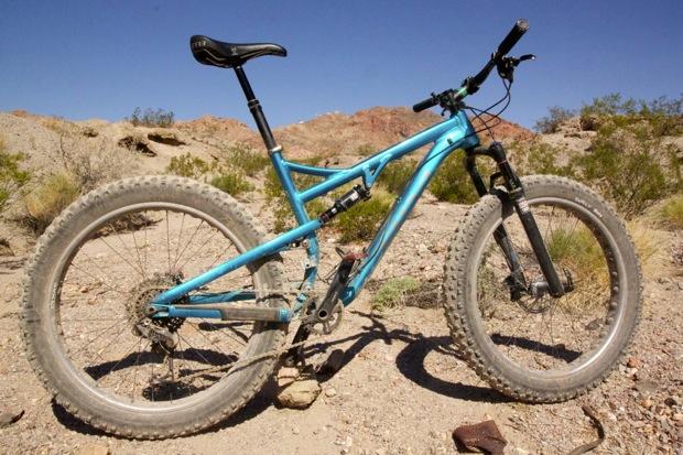 Salsa Bucksaw: A blue mountain bike is positioned on a rocky, desert terrain with scattered vegetation in the background and a clear blue sky. The bike features wide tires and a sturdy frame, designed for off-road riding.