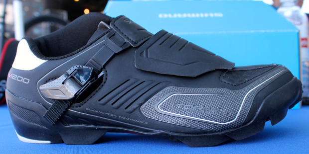 A close-up image of a black cycling shoe, featuring a sleek design with ridged details. The shoe has a dual strap closure system and a rigid sole suitable for performance cycling. In the background, there is a blue Shimano box partially visible. The shoe is displayed on a blue table surface.