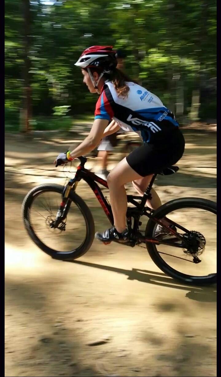 Trek Fuel EX 9: A cyclist wearing a helmet and a colorful jersey rides a mountain bike on a dirt trail, surrounded by greenery. The motion blur indicates speed as the rider navigates the path.