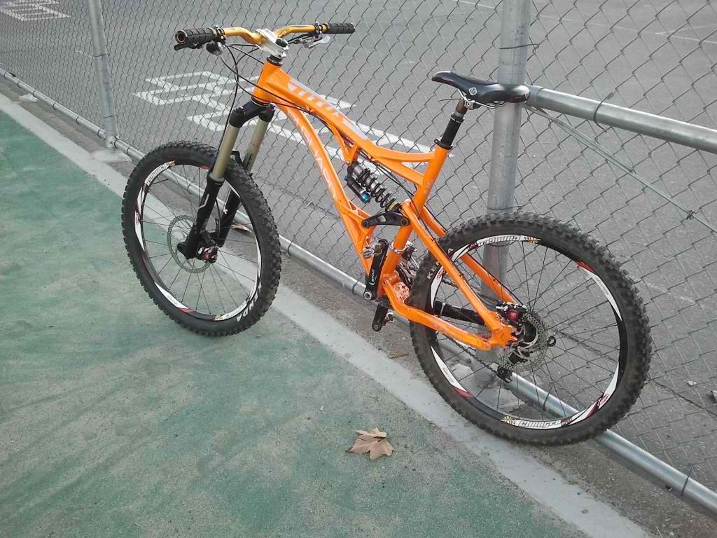 Titus El Guapo: A vibrant orange mountain bike leaning against a chain-link fence, with thick tires and visible suspension components. The bike is positioned on a green surface, indicating a cycling area or park. A fallen leaf is on the ground near the bike.