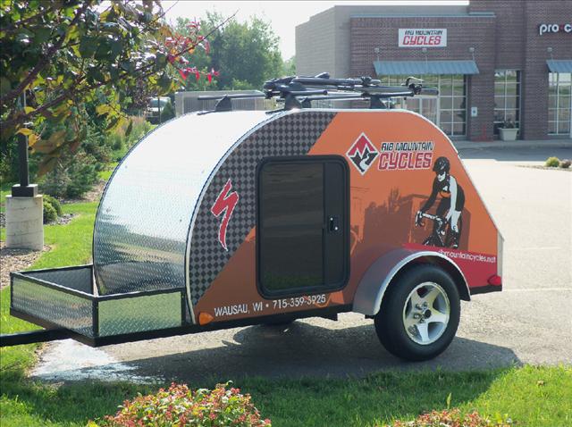 A small, rounded trailer with a checkered pattern and bright orange graphics, featuring the text "Air Mountain Cycles" and an illustration of a cyclist. The trailer has a black door and is parked in front of a commercial building surrounded by greenery.