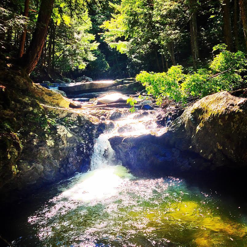 A tranquil waterfall flowing over rocks in a lush green forest, with sunlight filtering through the trees and creating dappled light on the water