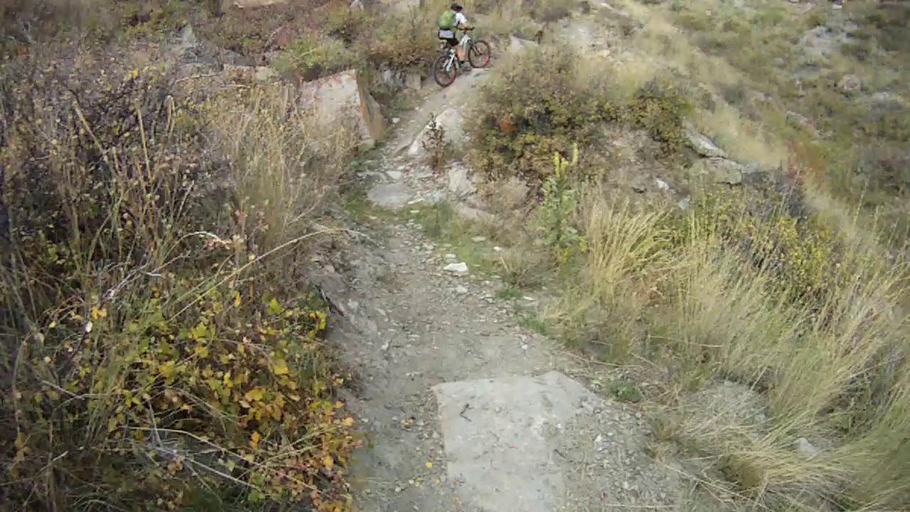 A person riding a mountain bike along a rocky trail surrounded by tall grass and vegetation. The terrain includes uneven ground with a mix of dirt and small stones. The scene showcases an outdoor adventure in a natural landscape. New Video Game/Yeah Kid mountain bike trail.